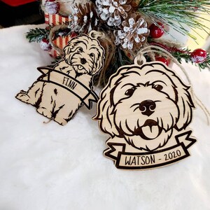 Op de afbeelding: Twee houten hondenornamenten met gegraveerde details. E&eacute;n ornament leest "FINN" op een banner, de andere "WATSON - 2020". Beide zijn lichtbruin met een natuurlijke houtnerf en hebben touw om op te hangen. Kerstversiering.