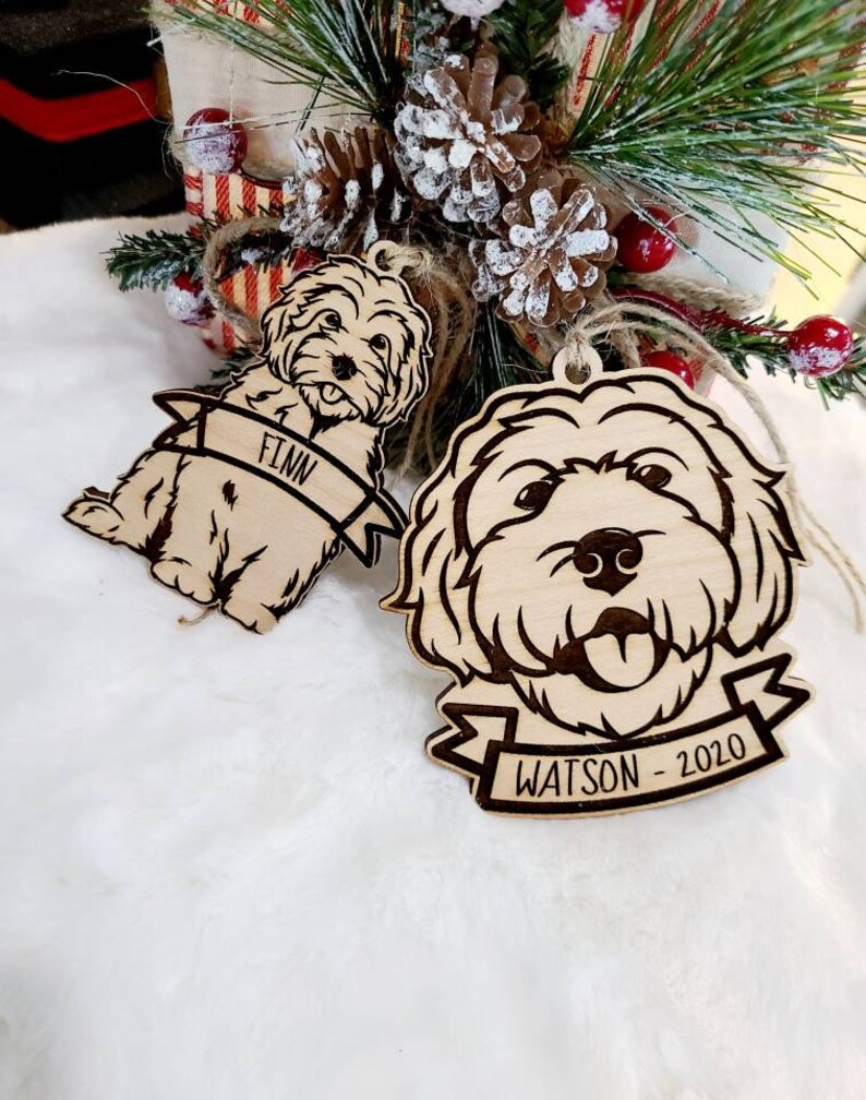 Op de afbeelding: Twee houten hondenornamenten met gegraveerde details. E&eacute;n ornament leest "FINN" op een banner, de andere "WATSON - 2020". Beide zijn lichtbruin met een natuurlijke houtnerf en hebben touw om op te hangen. Kerstversiering.