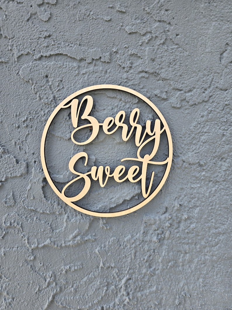 May include: A wooden circle with the words "Berry Sweet" written in cursive on it. The circle is a light brown color and is hanging on a gray wall.