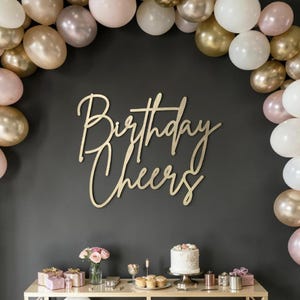 May include: A gold-colored wooden sign that reads "Birthday Cheers" is mounted on a dark gray wall. The sign is surrounded by a balloon arch in shades of gold, white, and pink. A table displays gifts, a cake, and cupcakes.