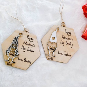 May include: Two hexagonal wooden ornaments featuring a gnome design. Each ornament depicts a white gnome holding a heart with the text "xoxo" on it. The text on the ornaments reads "Happy Valentine's Day Joshua Love, Brinley 2021" and "Happy Valentine's Day Brinley Love, Joshua 2021".