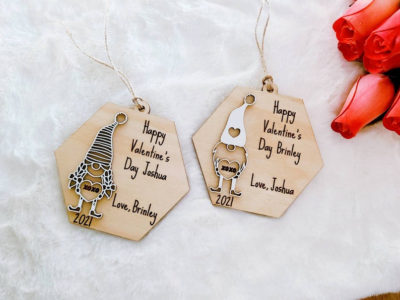 May include: Two hexagonal wooden ornaments featuring a gnome design. Each ornament depicts a white gnome holding a heart with the text "xoxo" on it. The text on the ornaments reads "Happy Valentine's Day Joshua Love, Brinley 2021" and "Happy Valentine's Day Brinley Love, Joshua 2021".