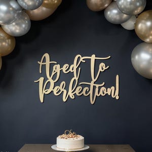 May include: Wooden sign reading 'Aged To Perfection!' in a stylish script, mounted on a dark wall. A cake with a gold topper is below, with gold and silver balloons in the background. A celebratory theme.