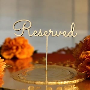 May include: A wooden cake topper with the word "Reserved" in elegant cursive script. The topper is set on a gold-colored base, surrounded by orange flowers. The overall theme suggests a wedding or event.