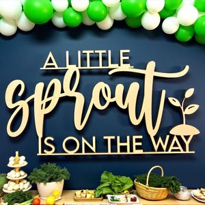 以下が含まれることがあります： 「A Little Sprout Is On The Way」と書かれた木製の看板が、緑と白の風船で飾られた濃い青色の壁に飾られています。 下のテーブルには食べ物と緑が飾られています。