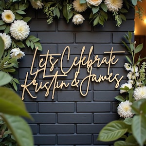 Puede incluir: Letrero de madera con la frase "Let's Celebrate Kristin & James" en elegante escritura cursiva. El letrero est&aacute; colocado sobre una pared de ladrillos gris oscuro, rodeado de flores blancas y follaje verde, creando un ambiente festivo.