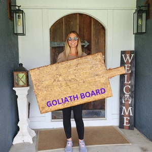Puede incluir: Una tabla de cortar de madera rectangular grande con un asa, con la etiqueta "GOLIATH BOARD" en texto morado. La tabla tiene un acabado de vetas de madera natural. La persona est&aacute; de pie frente a una puerta de madera.