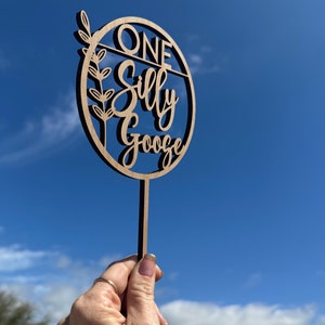 May include: A wooden cake topper with the text "ONE Silly Goose" in a circle with a leafy branch design. The topper is held in a hand against a blue sky background.