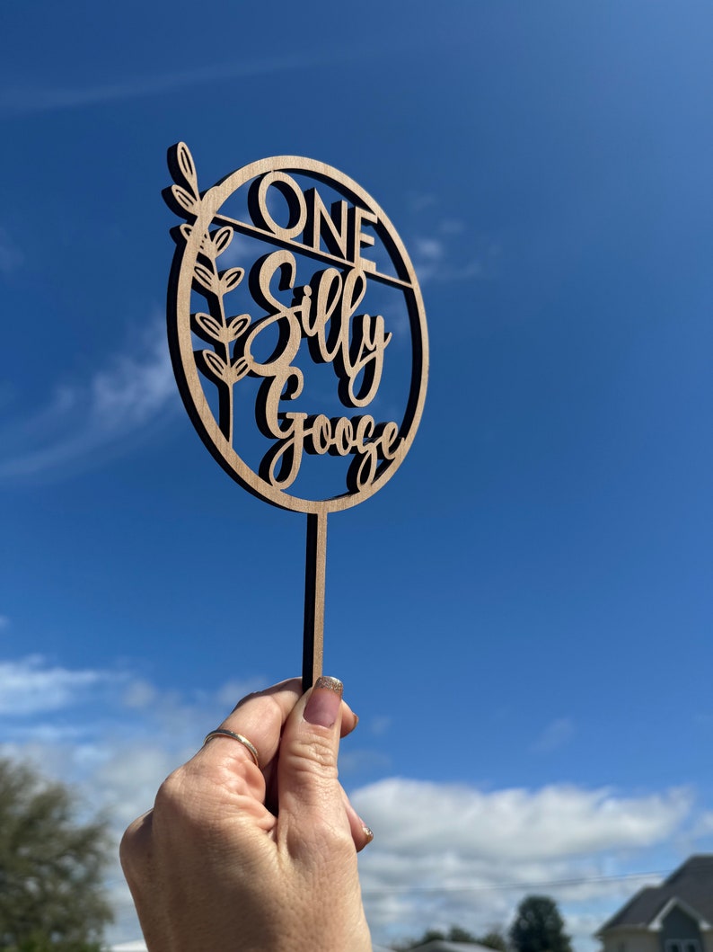 May include: A wooden cake topper with the text "ONE Silly Goose" in a circle with a leafy branch design. The topper is held in a hand against a blue sky background.