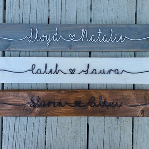 May include: Three wooden signs with names and a heart symbol written in wire. The top sign is gray, the middle sign is white, and the bottom sign is brown.