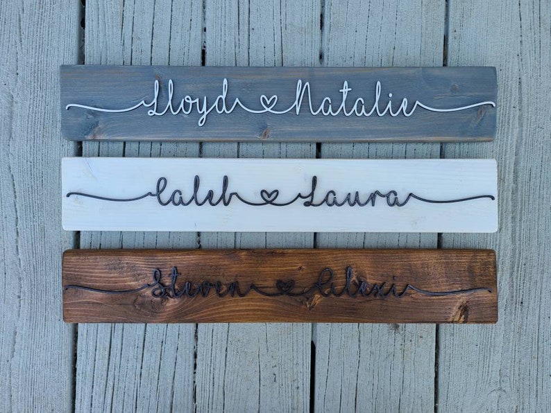 May include: Three wooden signs with names and a heart symbol written in wire. The top sign is gray, the middle sign is white, and the bottom sign is brown.