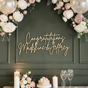 May include: A gold-toned script sign reading Congratulations Madeleine & Jeffrey mounted on a dark green panelled wall. The display is framed by white and pink floral arrangements and two metallic balloons, with candles and wine glasses below.