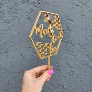 May include: A wooden cake topper in the shape of a hexagon with the words "Mama to Bee" and a bee design. The topper is on a stick and is held by a hand.