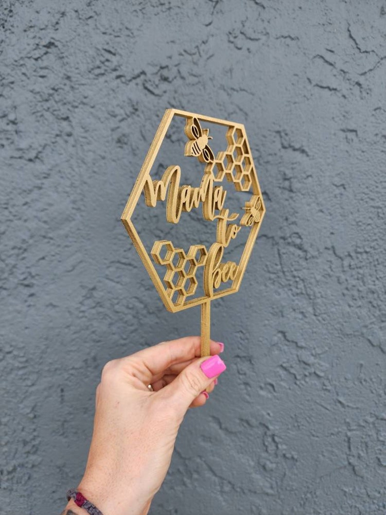 May include: A wooden cake topper in the shape of a hexagon with the words "Mama to Bee" and a bee design. The topper is on a stick and is held by a hand.