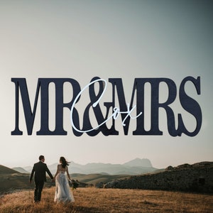 May include: A dark blue wooden sign with the words "MR & MRS" and the last name "Cox" in a cursive font. The sign is displayed above a couple walking hand-in-hand in a field, with mountains in the background. Wedding decor.