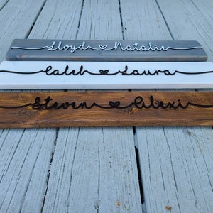 May include: Three wooden signs with names and a heart symbol carved into them. The top sign is gray, the middle sign is white, and the bottom sign is brown. The names on the signs are "Lloyd & Natalie", "Caleb & Laura", and "Steven & Alexi".
