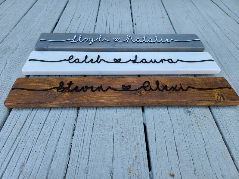 May include: Three wooden signs with names and a heart symbol carved into them. The top sign is gray, the middle sign is white, and the bottom sign is brown. The names on the signs are "Lloyd & Natalie", "Caleb & Laura", and "Steven & Alexi".