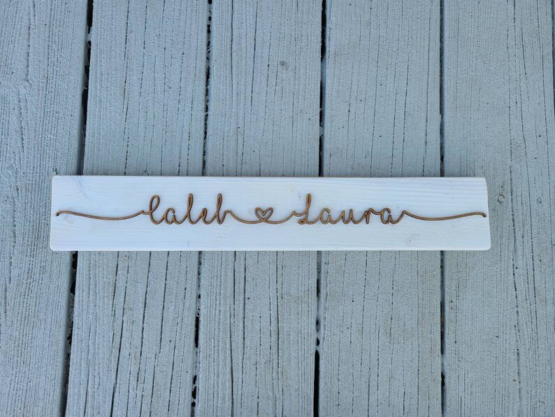 May include: White wooden sign with the names "Caleb" and "Laura" written in cursive with a heart symbol between them.