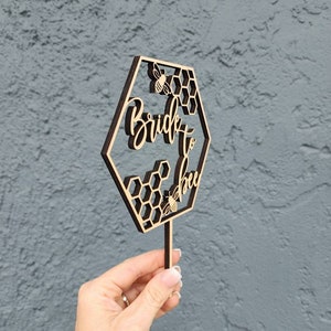 May include: Wooden cake topper with a hexagonal design featuring a honeycomb pattern and the text "Bride to Bee" with a bee illustration.