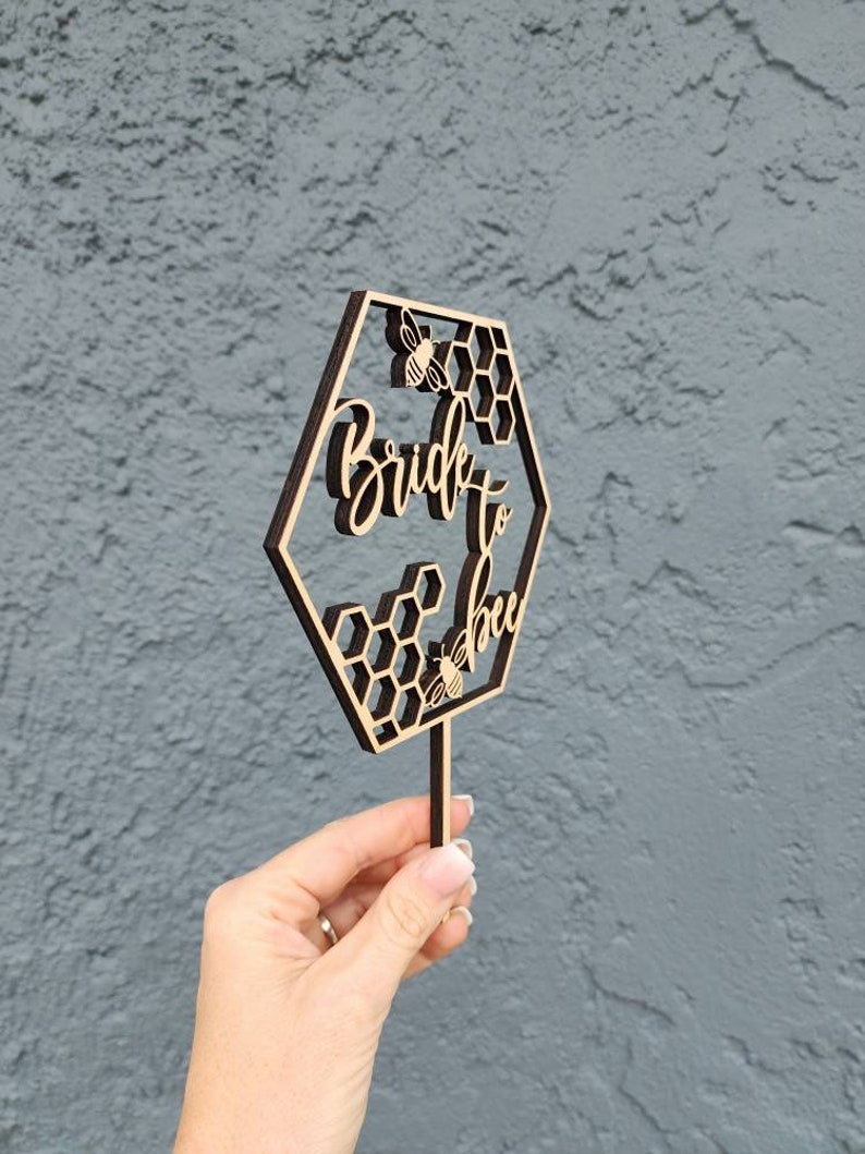 May include: Wooden cake topper with a hexagonal design featuring a honeycomb pattern and the text "Bride to Bee" with a bee illustration.
