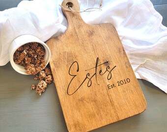 May include: A wooden cutting board with a handle, featuring the engraved text "Estes Est. 2010". The board has a natural wood grain and a light brown color.