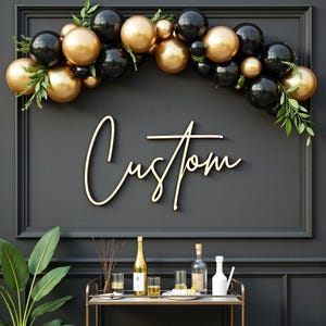 Op de afbeelding: Een zwarte muur met een goudkleurig "Custom" bord. Boven het bord een ballonnenboog van gouden en zwarte ballonnen. Een barwagen met flessen en glazen staat eronder. Groen is verspreid over de ballonnenboog.