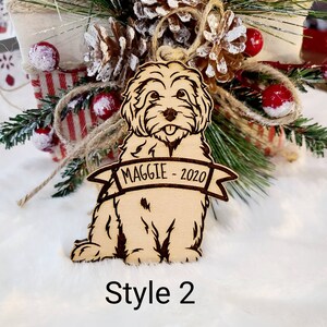 Op de afbeelding: Lichtbruine houten hondenornament met de naam "MAGGIE - 2020" gegraveerd op een banner. De hond wordt zittend afgebeeld met zijn tong uit. Het ornament heeft donkerbruine ge&euml;tste details en is omgeven door kerstversieringen.