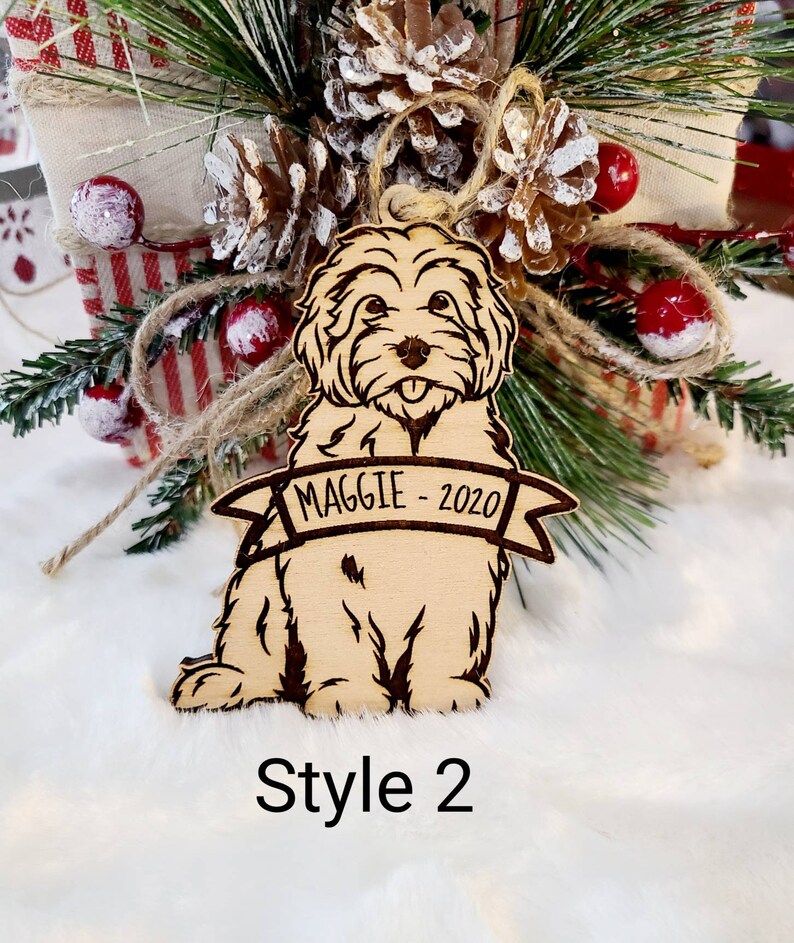 Op de afbeelding: Lichtbruine houten hondenornament met de naam "MAGGIE - 2020" gegraveerd op een banner. De hond wordt zittend afgebeeld met zijn tong uit. Het ornament heeft donkerbruine ge&euml;tste details en is omgeven door kerstversieringen.