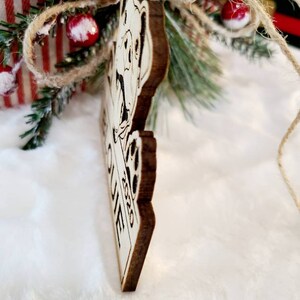 K&ouml;nnte beinhalten: Weihnachtsbaumschmuck aus Holz mit einem Hundedesign und dem Text "Our Home 2020". Der Anh&auml;nger ist braun mit einer hellen Vorderseite und wird an einer Schnur aufgeh&auml;ngt. Der Hintergrund umfasst Weihnachtsdekorationen.
