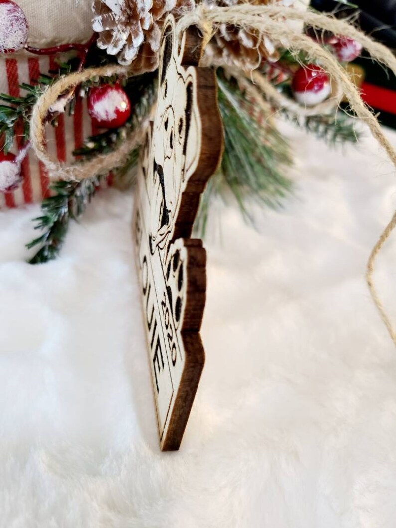 K&ouml;nnte beinhalten: Weihnachtsbaumschmuck aus Holz mit einem Hundedesign und dem Text "Our Home 2020". Der Anh&auml;nger ist braun mit einer hellen Vorderseite und wird an einer Schnur aufgeh&auml;ngt. Der Hintergrund umfasst Weihnachtsdekorationen.