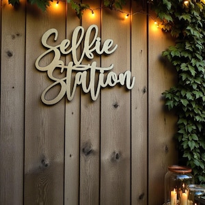 Puede incluir: Un letrero de madera con la inscripci&oacute;n "Selfie Station" en letra cursiva est&aacute; montado en una valla de madera. El letrero est&aacute; rodeado de vegetaci&oacute;n y luces de cadena. Velas en tarros de cristal sobre una mesa en la esquina inferior derecha.