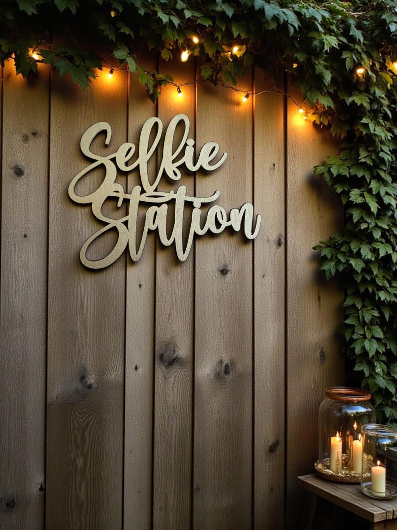 Puede incluir: Un letrero de madera con la inscripci&oacute;n "Selfie Station" en letra cursiva est&aacute; montado en una valla de madera. El letrero est&aacute; rodeado de vegetaci&oacute;n y luces de cadena. Velas en tarros de cristal sobre una mesa en la esquina inferior derecha.