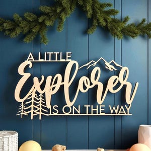 May include: Wooden sign with the text "A Little Explorer Is On The Way" in a cursive font, featuring mountain and tree illustrations. The sign is mounted on a blue wall, with a green garland above and decorative items below.