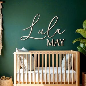 Puede incluir: Una cuna de madera con un colch&oacute;n blanco y dos almohadas grises est&aacute; frente a una pared verde oscuro. El nombre "Lula MAY" se muestra en letras cursivas de madera sobre la cuna. Una cesta tejida y una planta en maceta est&aacute;n a ambos lados de la cuna.