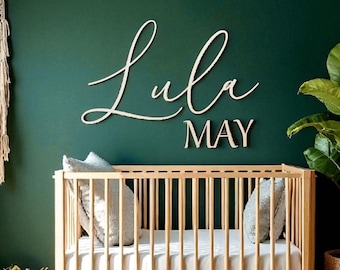 May include: A wooden crib with a white mattress and two gray pillows is in front of a dark green wall. The name "Lula MAY" is displayed in wooden cursive letters above the crib. A woven basket and a potted plant are on either side of the crib.