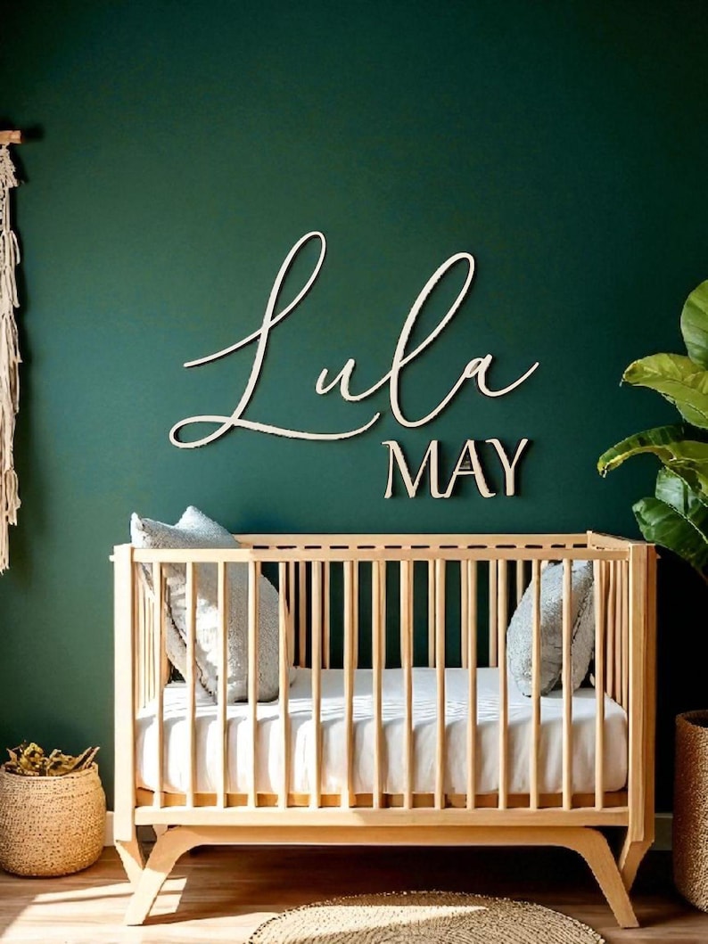 Puede incluir: Una cuna de madera con un colch&oacute;n blanco y dos almohadas grises est&aacute; frente a una pared verde oscuro. El nombre "Lula MAY" se muestra en letras cursivas de madera sobre la cuna. Una cesta tejida y una planta en maceta est&aacute;n a ambos lados de la cuna.