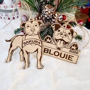K&ouml;nnte beinhalten: Zwei Hundeornamente aus Holz. Eines ist ein ganzer K&ouml;rper eines Hundes mit dem Namen "Sakura" auf einem Banner. Das andere ist ein Hundekopf mit dem Namen "Blouie" und dem Jahr "2020" auf einem Banner. Beide h&auml;ngen mit Bindfaden.
