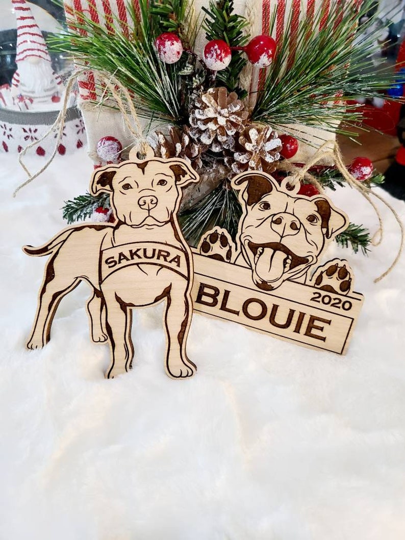 K&ouml;nnte beinhalten: Zwei Hundeornamente aus Holz. Eines ist ein ganzer K&ouml;rper eines Hundes mit dem Namen "Sakura" auf einem Banner. Das andere ist ein Hundekopf mit dem Namen "Blouie" und dem Jahr "2020" auf einem Banner. Beide h&auml;ngen mit Bindfaden.