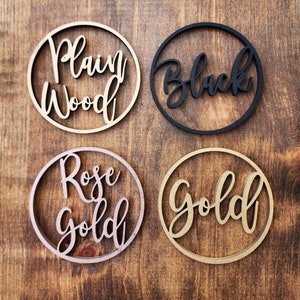 May include: Four wooden circles with different finishes: plain wood, black, rose gold, and gold. Each circle has a word cut out in the center.