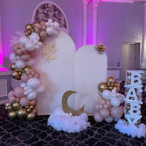 Puede incluir: Decoraci&oacute;n de baby shower con un tel&oacute;n de fondo blanco y letras doradas que dicen "A Little Love From Above". La decoraci&oacute;n incluye arcos de globos en blanco, dorado y rosa, una luna creciente dorada y un letrero de "BABY".
