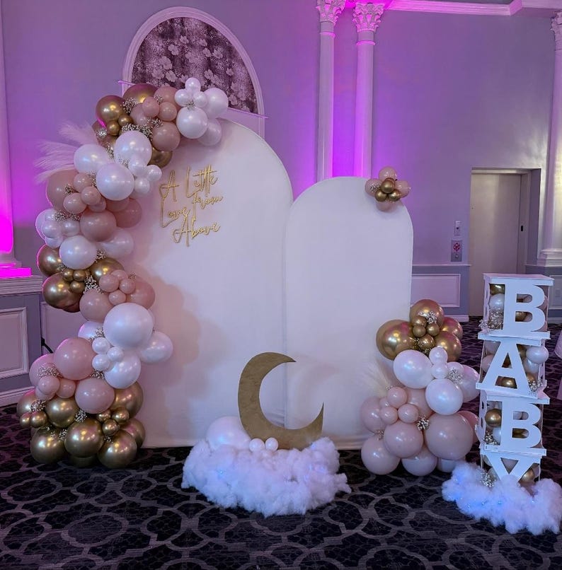 Puede incluir: Decoraci&oacute;n de baby shower con un tel&oacute;n de fondo blanco y letras doradas que dicen "A Little Love From Above". La decoraci&oacute;n incluye arcos de globos en blanco, dorado y rosa, una luna creciente dorada y un letrero de "BABY".