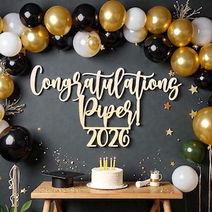 以下が含まれることがあります： 卒業のお祝いのシーン。木製の看板には「Congratulations Piper! 2026」と書かれています。シーンには、金、黒、白の風船、ろうそくのついたケーキ、卒業帽子、小さなテーブルが含まれています。