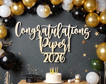 Puede incluir: Escena de celebración de graduación con un letrero de madera que dice "¡Felicidades Piper! 2026". La escena incluye globos dorados, negros y blancos, un pastel con velas, un birrete y una pequeña mesa.