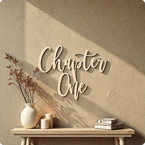 May include: Wooden wall art spelling "Chapter One" in a cursive font. The art is mounted on a beige wall, with a shadow cast by sunlight. A white vase with dried flowers, candles, and books are on a wooden shelf below.