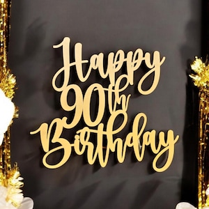 May include: A gold-coloured wooden cake topper with the words "Happy 90th Birthday" in elegant script. The topper is set against a dark background, with gold streamers and white flowers in the corners.