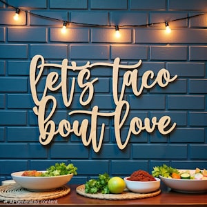 Puede incluir: Letrero de madera con la frase "let's taco bout love" en escritura cursiva, montado en una pared de ladrillos azules. La escena incluye luces de cadena y cuencos de comida, sugiriendo un ambiente festivo.