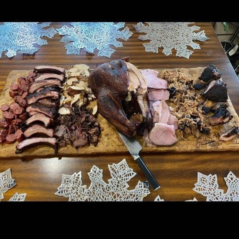 Puede incluir: Una tabla de servir de madera exhibe una variedad de carnes ahumadas. La disposici&oacute;n incluye costillas, salchichas, pavo, jam&oacute;n y cerdo desmenuzado. Un cuchillo para trinchar descansa sobre la tabla. La comida est&aacute; dispuesta sobre una mesa de madera con manteles individuales decorativos.