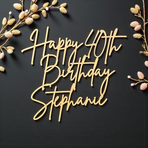 Puede incluir: Un adorno dorado para pastel con las palabras "Happy 40th Birthday Stephanie" en una fuente cursiva. El adorno est&aacute; sobre un fondo oscuro y decorado con detalles florales.