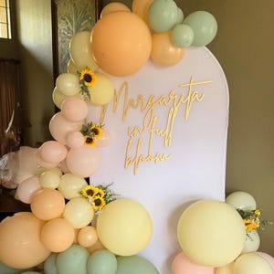 Puede incluir: Un tel&oacute;n de fondo para fiestas con un arco blanco y la inscripci&oacute;n dorada "Margarita in full bloom". Globos de colores pastel melocot&oacute;n, amarillo y verde, y girasoles decoran el arco. Los globos est&aacute;n dispuestos en racimos.
