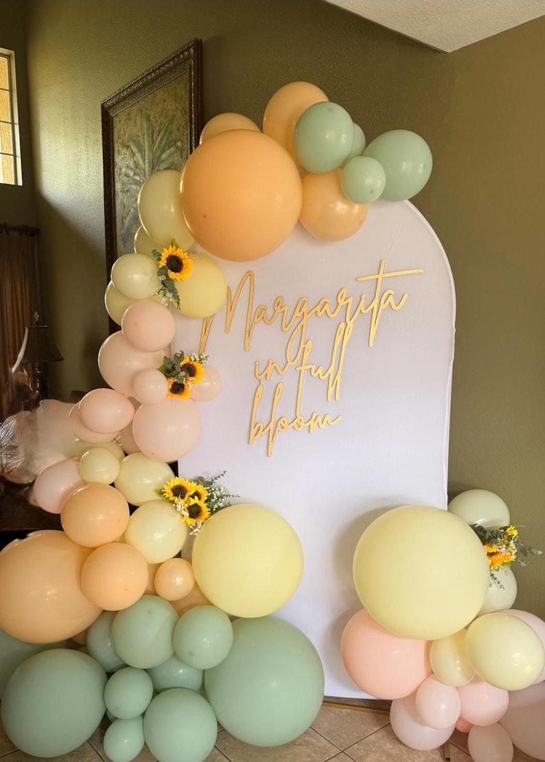 Puede incluir: Un tel&oacute;n de fondo para fiestas con un arco blanco y la inscripci&oacute;n dorada "Margarita in full bloom". Globos de colores pastel melocot&oacute;n, amarillo y verde, y girasoles decoran el arco. Los globos est&aacute;n dispuestos en racimos.